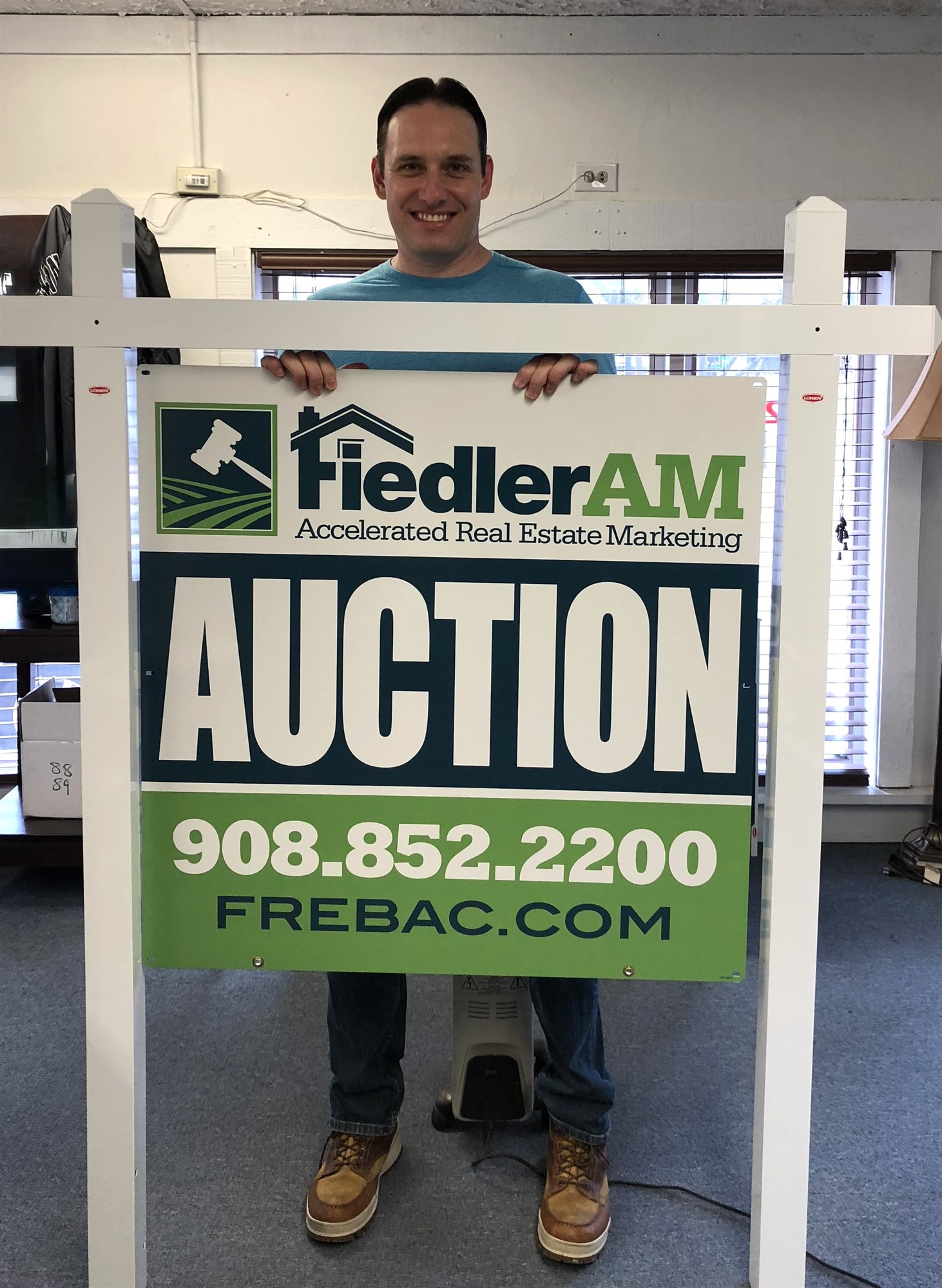 A younger Jeff Fiedler grins behind a FiedlerAM Accelerated Real Estate Marketing auction sign panel, held in its white post frame inside the office.