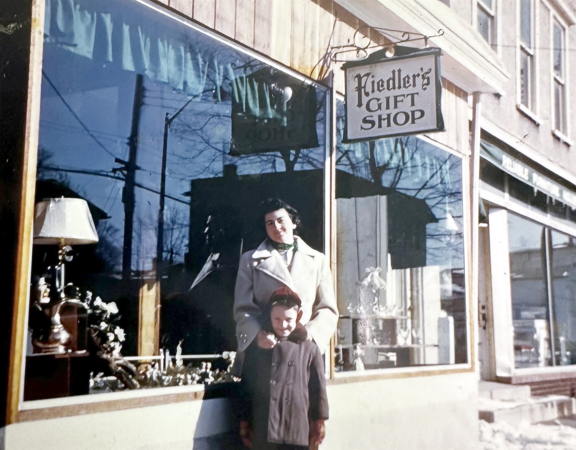 A woman in a light winter coat and a young boy stand on a sunny sidewalk in front of a storefront window; a hanging sign reads 'Fiedler's Gift Shop.'