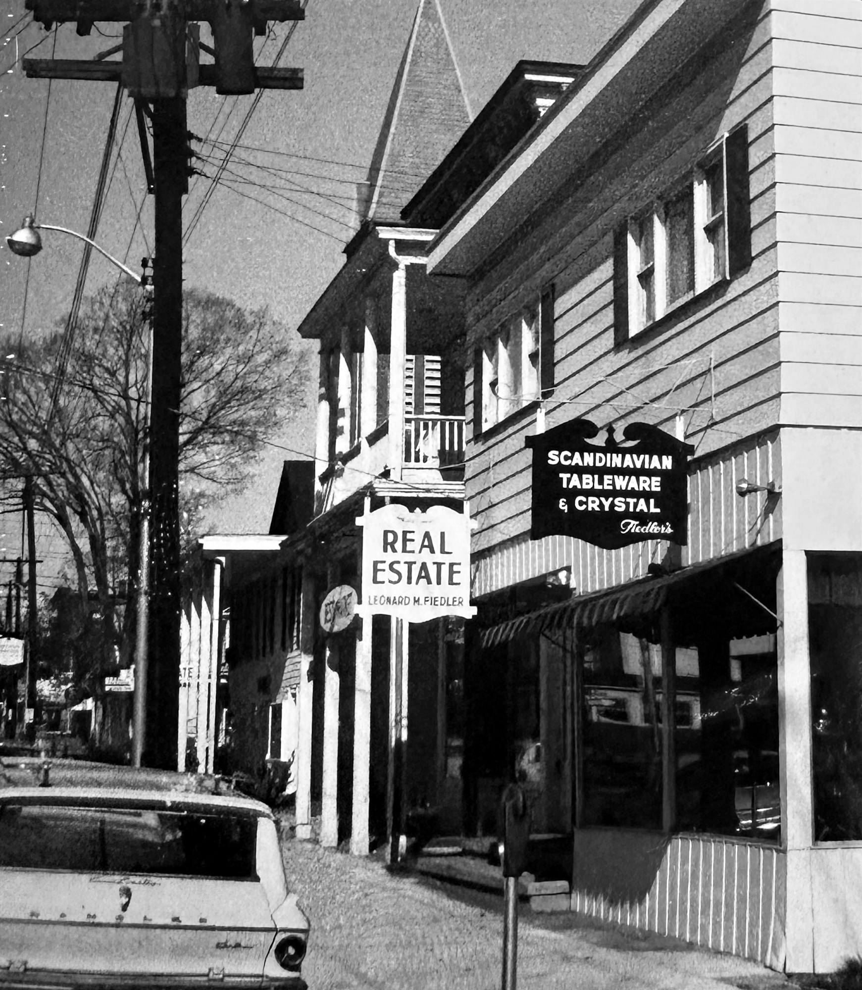 A Main Street building with two hanging signs — 'Real Estate, Leonard M. Fiedler' and 'Scandinavian Tableware & Crystal, Fiedler's' — and an old car parked at the curb.