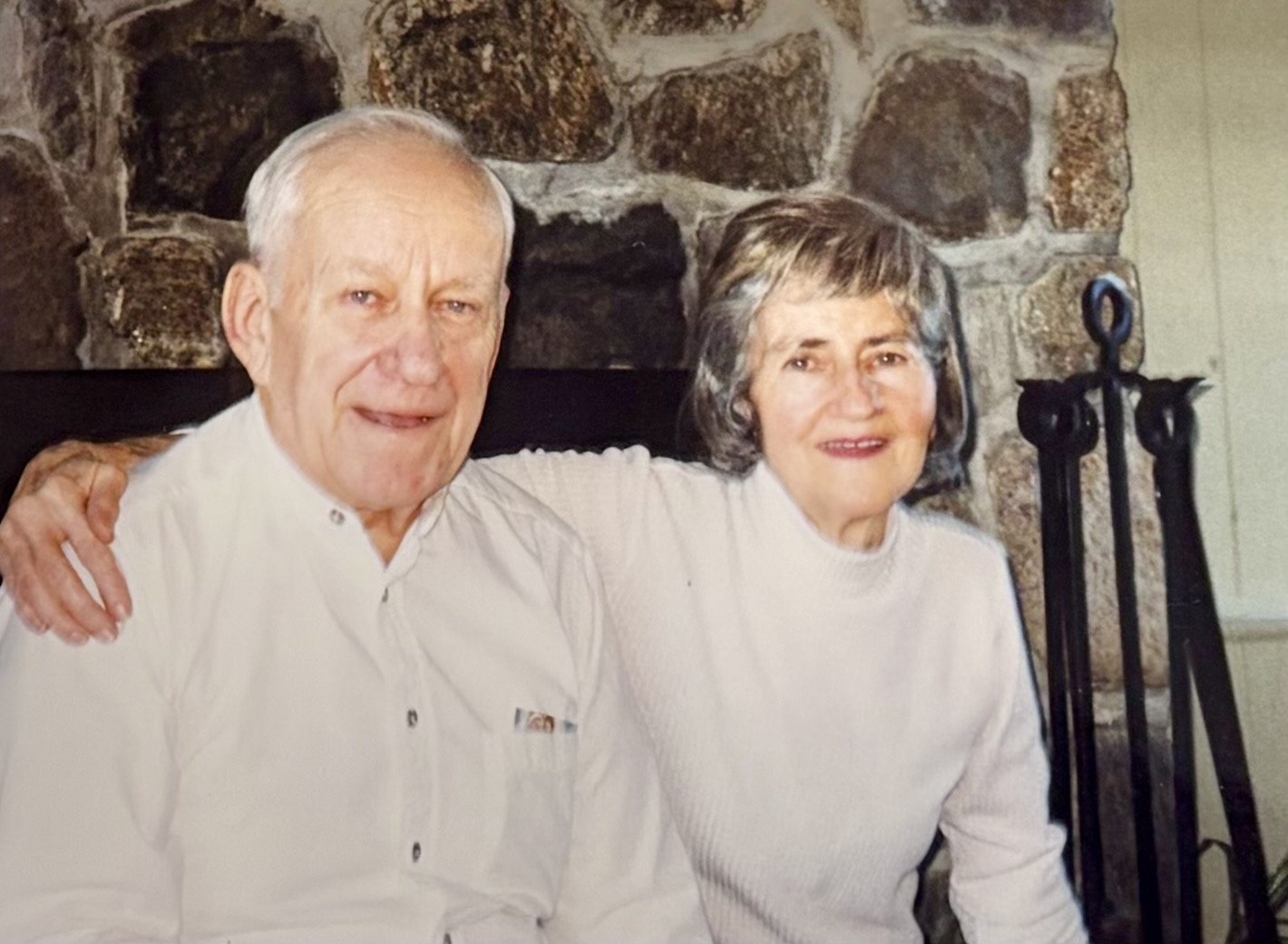 Helen and Leonard Fiedler in their later years, seated together in front of a stone fireplace — Leonard's arm around Helen, both in white, both smiling.