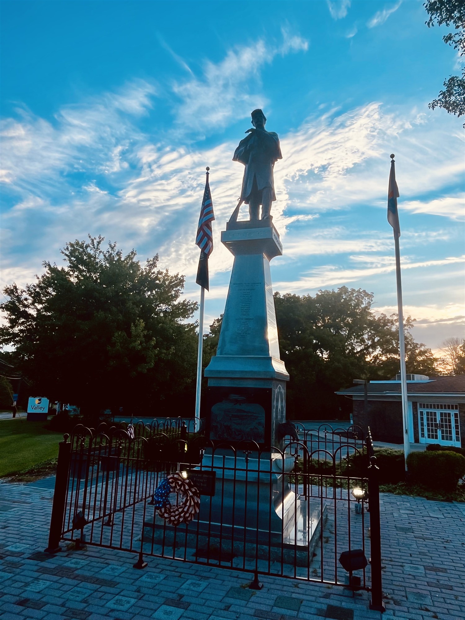 The Civil War soldier monument at evening — the statue silhouetted against a swirling blue sky, flanked by flags, a patriotic wreath on its iron fence.