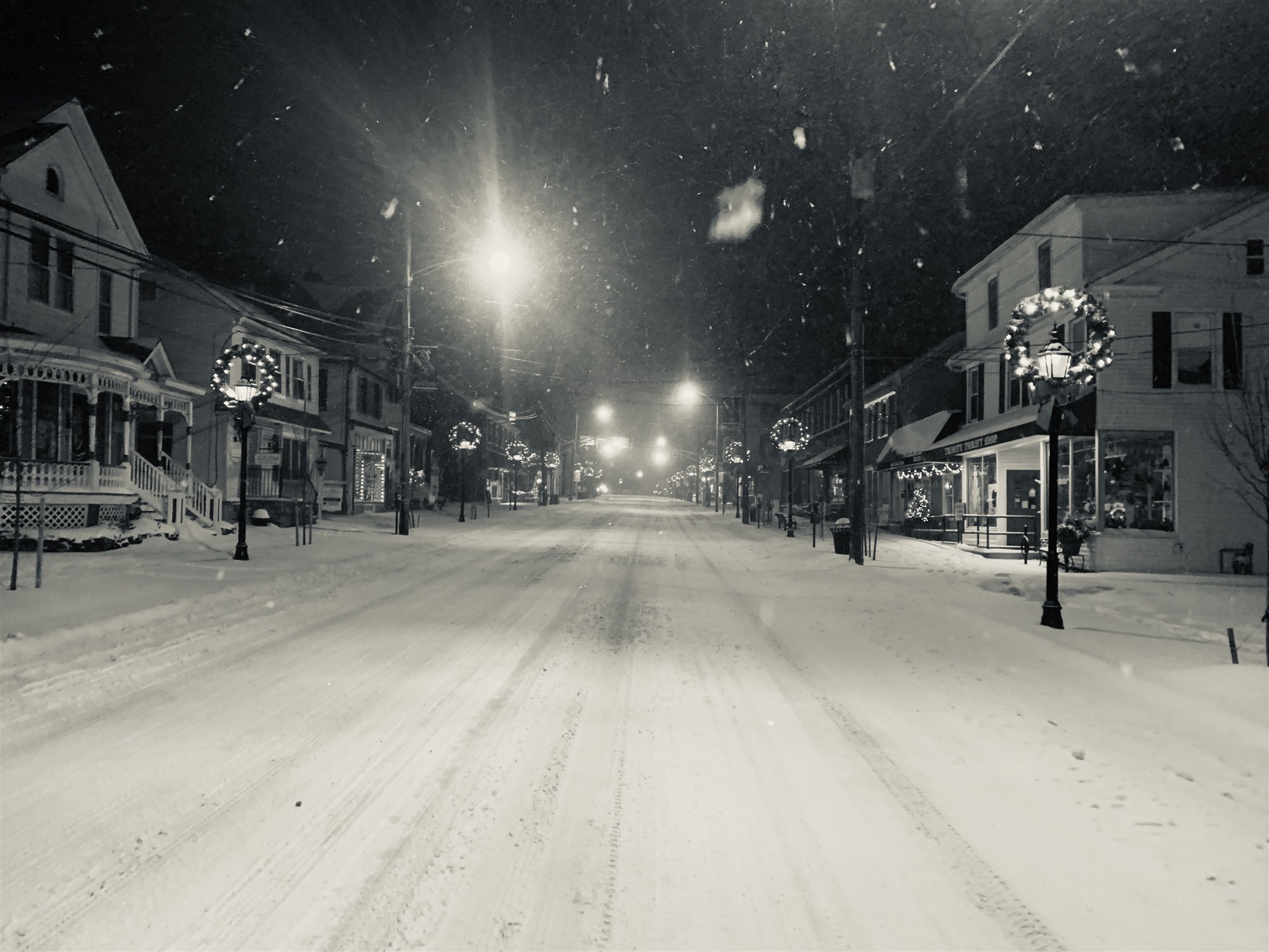 Snow-covered Main Street at night, empty and quiet in falling snow, Christmas wreaths glowing on the lampposts.