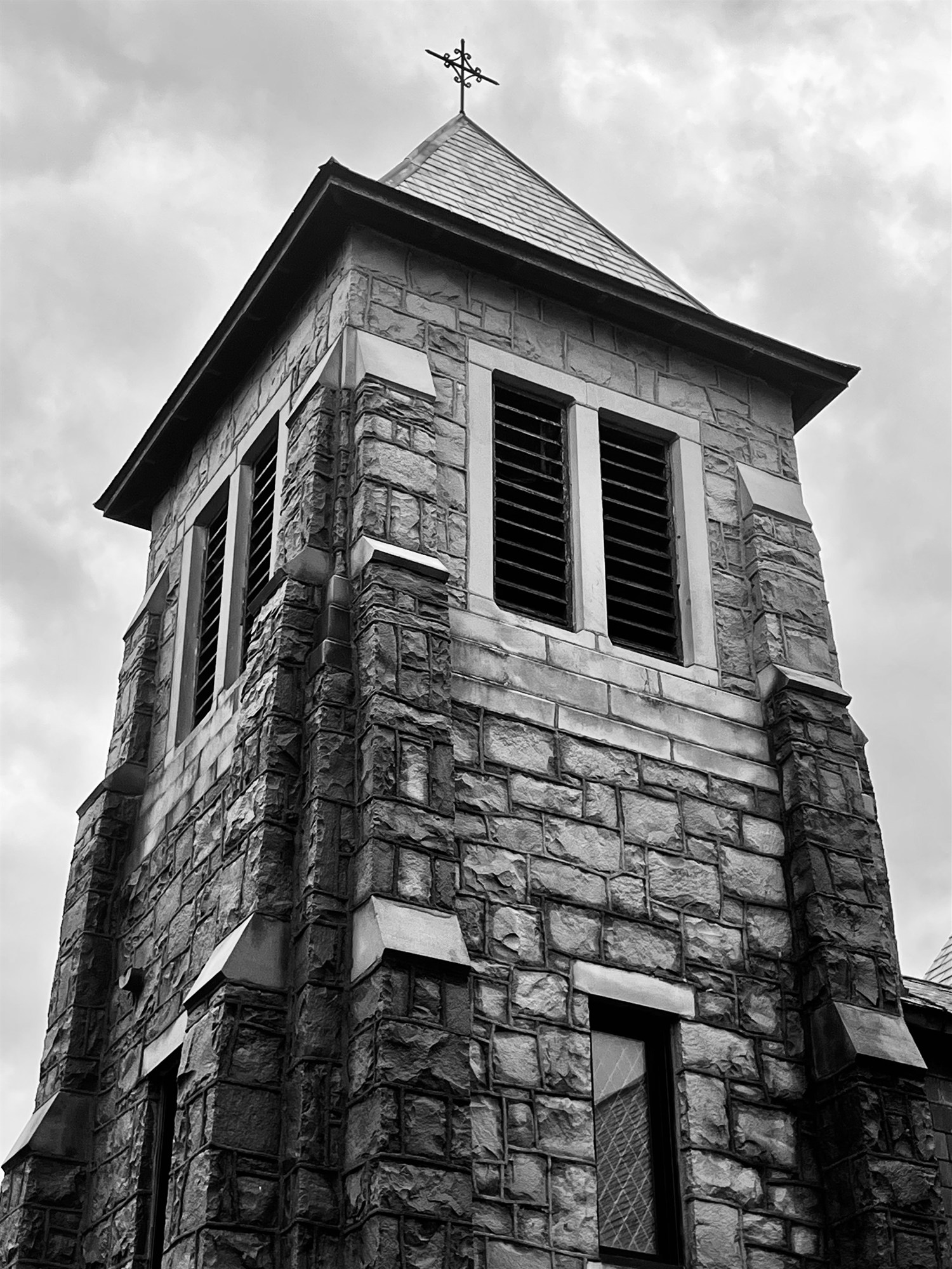 The rough-stone bell tower of St. Mary of Assumption, topped with a wrought-iron cross, against a gray sky.