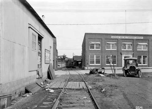 A historic black-and-white photograph of the American Saw Mill Machinery Company works: a two-story brick factory with the company name across it, a rail spur and a 1920s car in front.