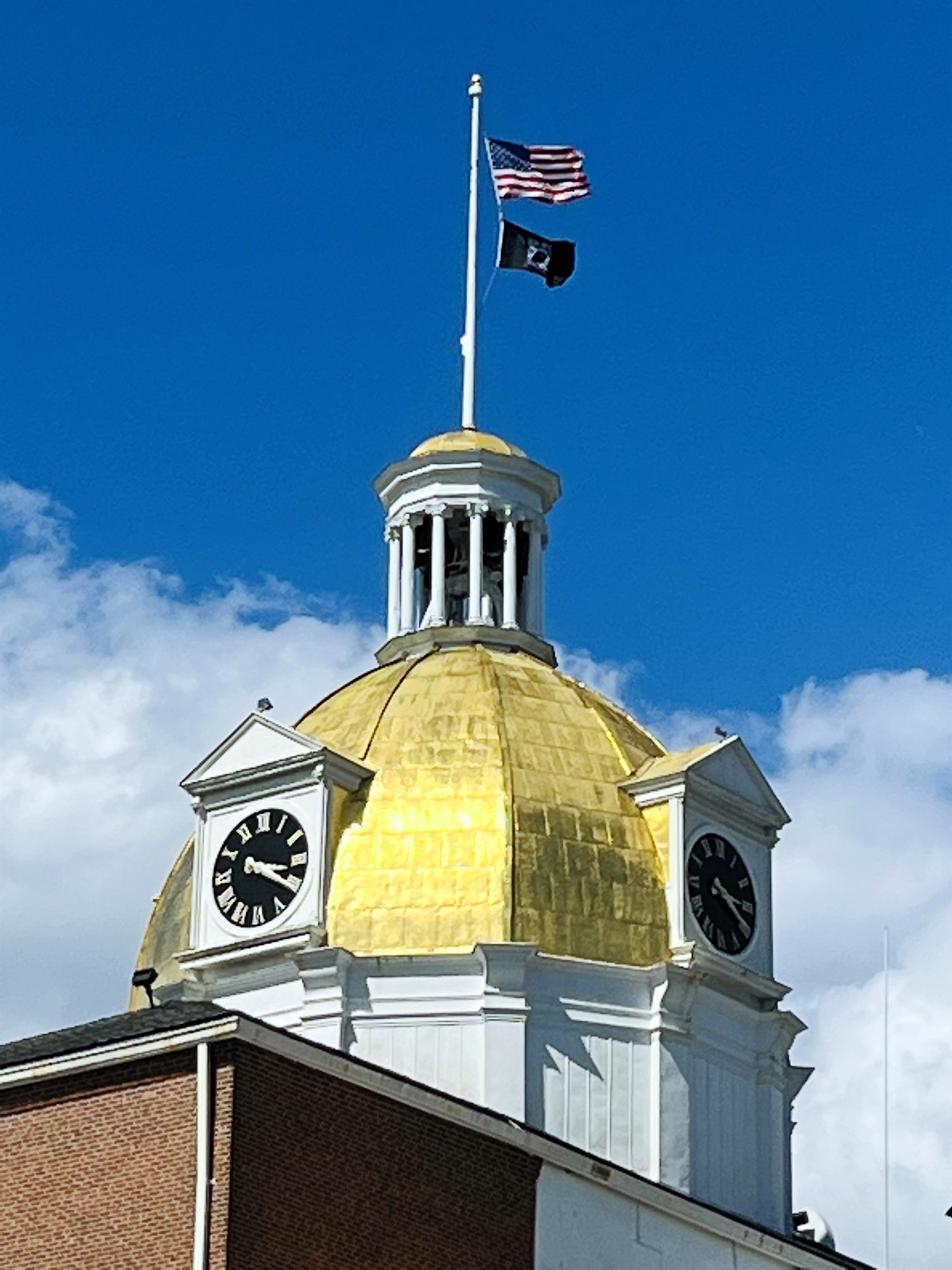A close view of the building's golden dome and white cupola, topped by an American flag and a POW/MIA flag and flanked by two black-faced clocks, against a blue sky.