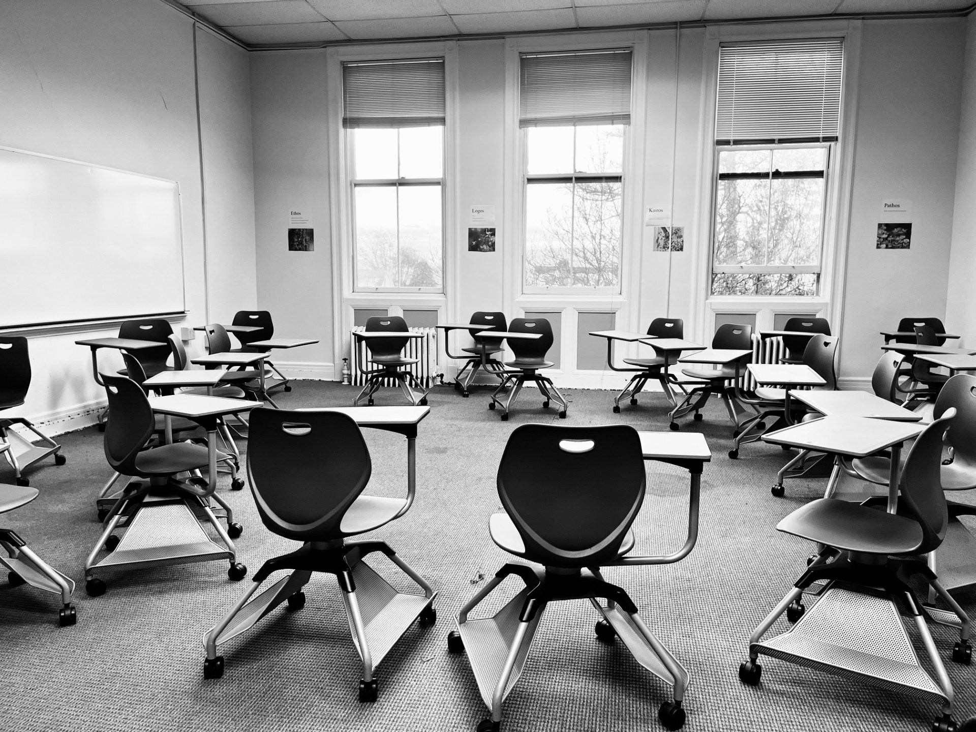 A black-and-white view of an old classroom in the main building: modern wheeled desks on a carpeted floor, tall windows, and rhetoric terms posted on the walls.