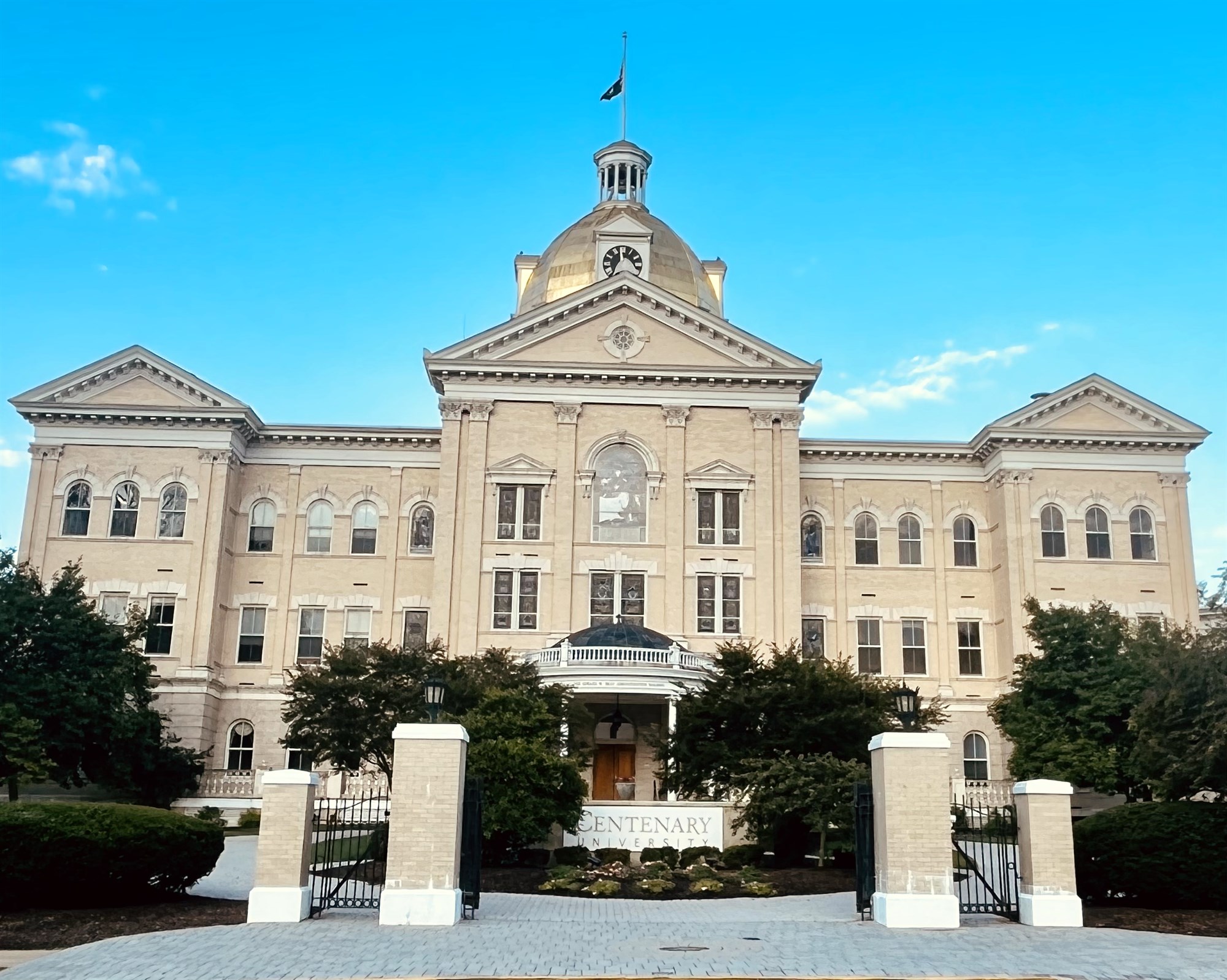 Centenary University's historic three-story main building with a golden dome and clock, seen in daylight from the front gate lettered 'Centenary University' under a blue sky.
