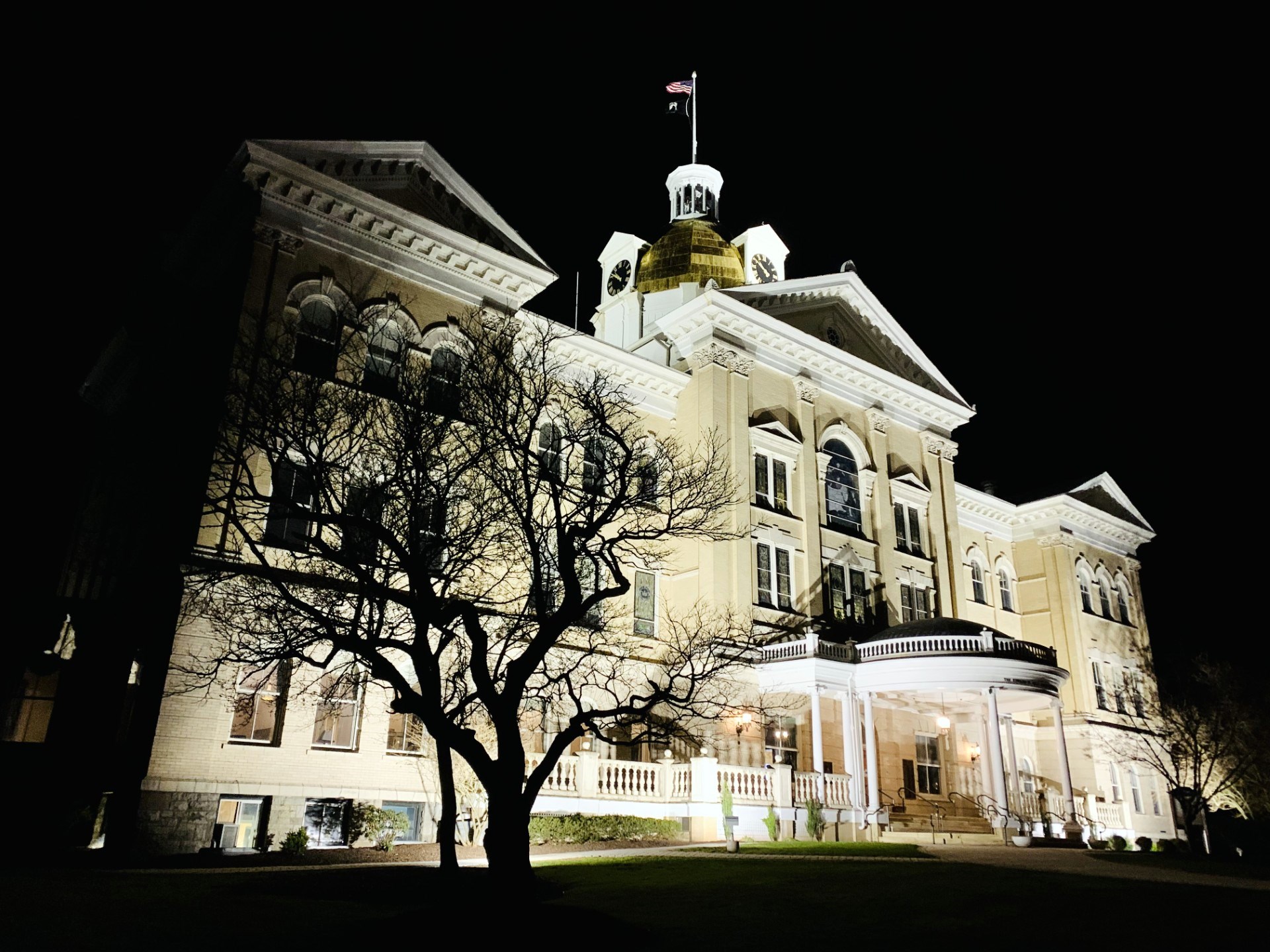 The Centenary main building lit up at night, its golden dome glowing, with a bare tree silhouetted in the foreground against a black sky.