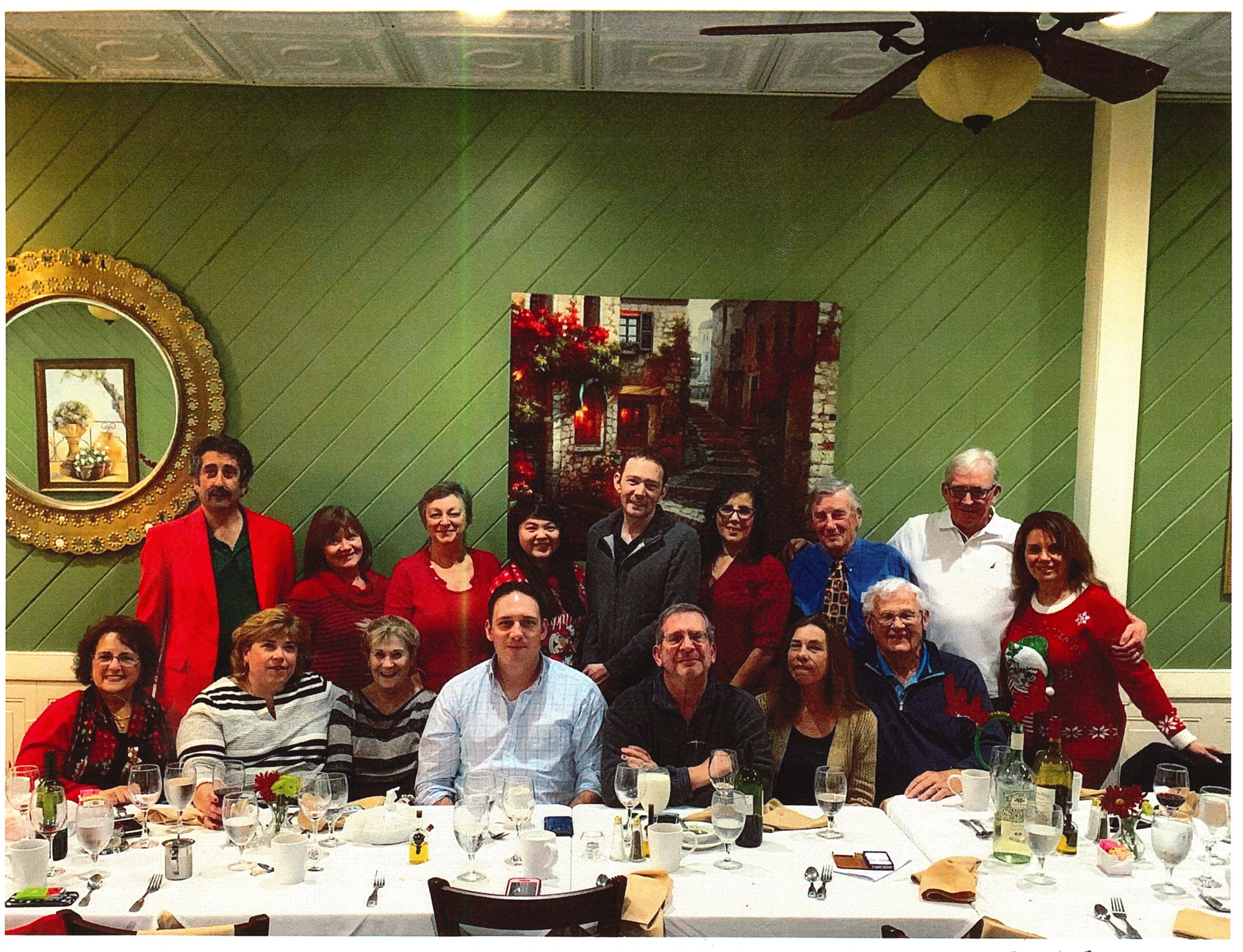 A long white table at the restaurant with sixteen team members seated and standing, most in red, under a tin ceiling beside a gold mirror.