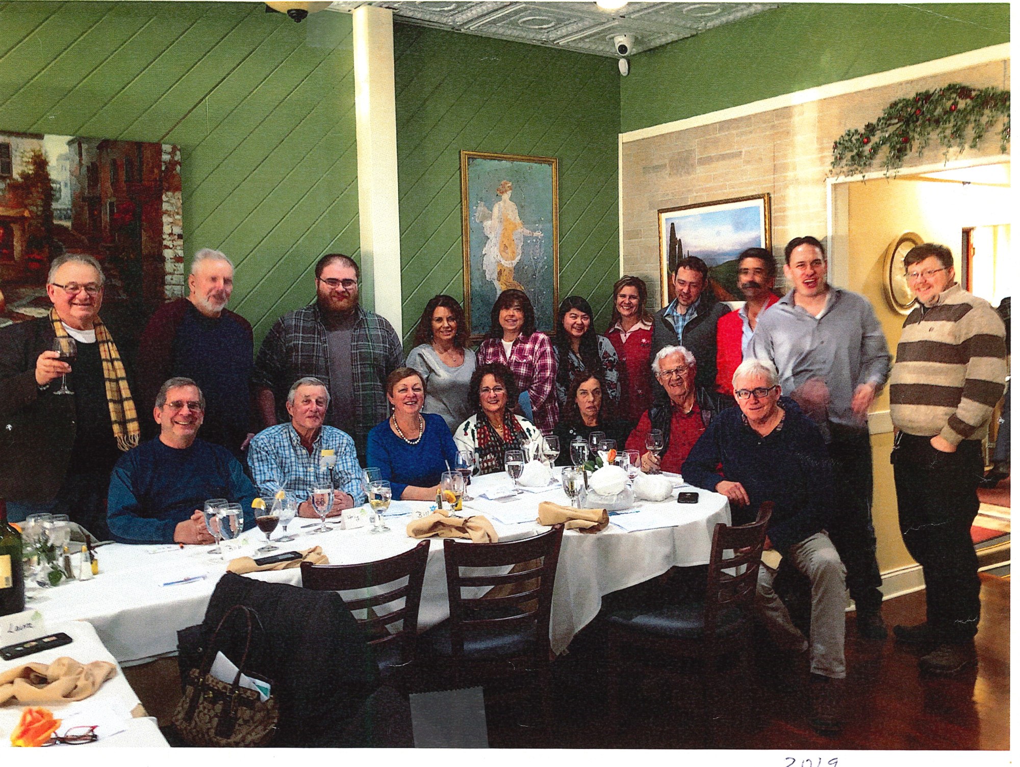 Eighteen team members seated and standing around white-clothed tables in the restaurant's corner room, name cards and wine glasses on the table.