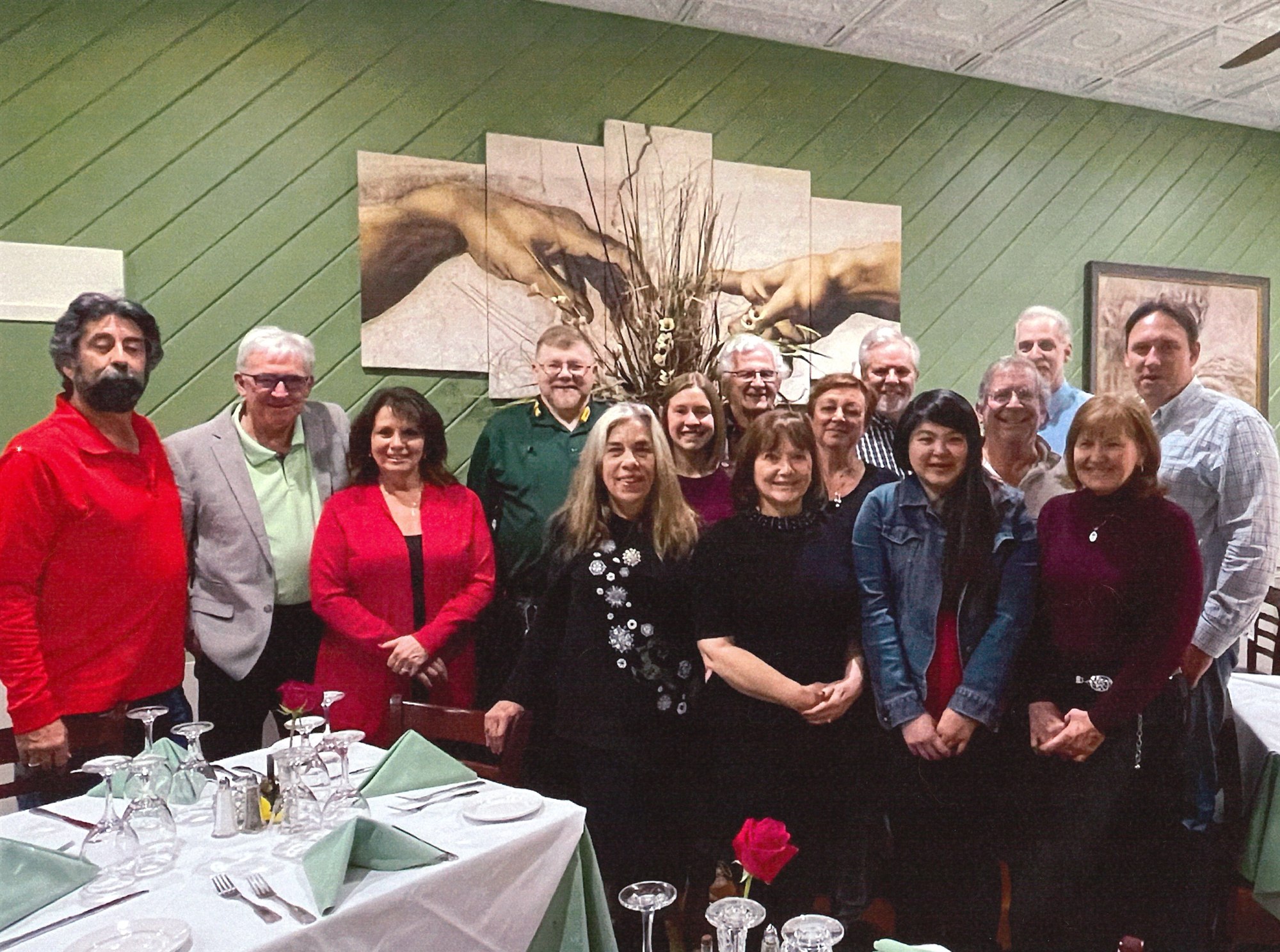 Fifteen team members standing together between set tables, in front of a wall-sized painting of two reaching hands on a green wall.