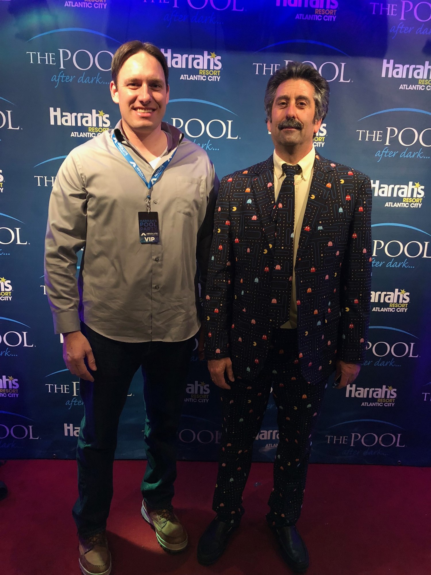 Two men at a step-and-repeat backdrop at Harrah's Atlantic City, one in a gray shirt with a VIP lanyard, the other in a Pac-Man patterned suit and tie.