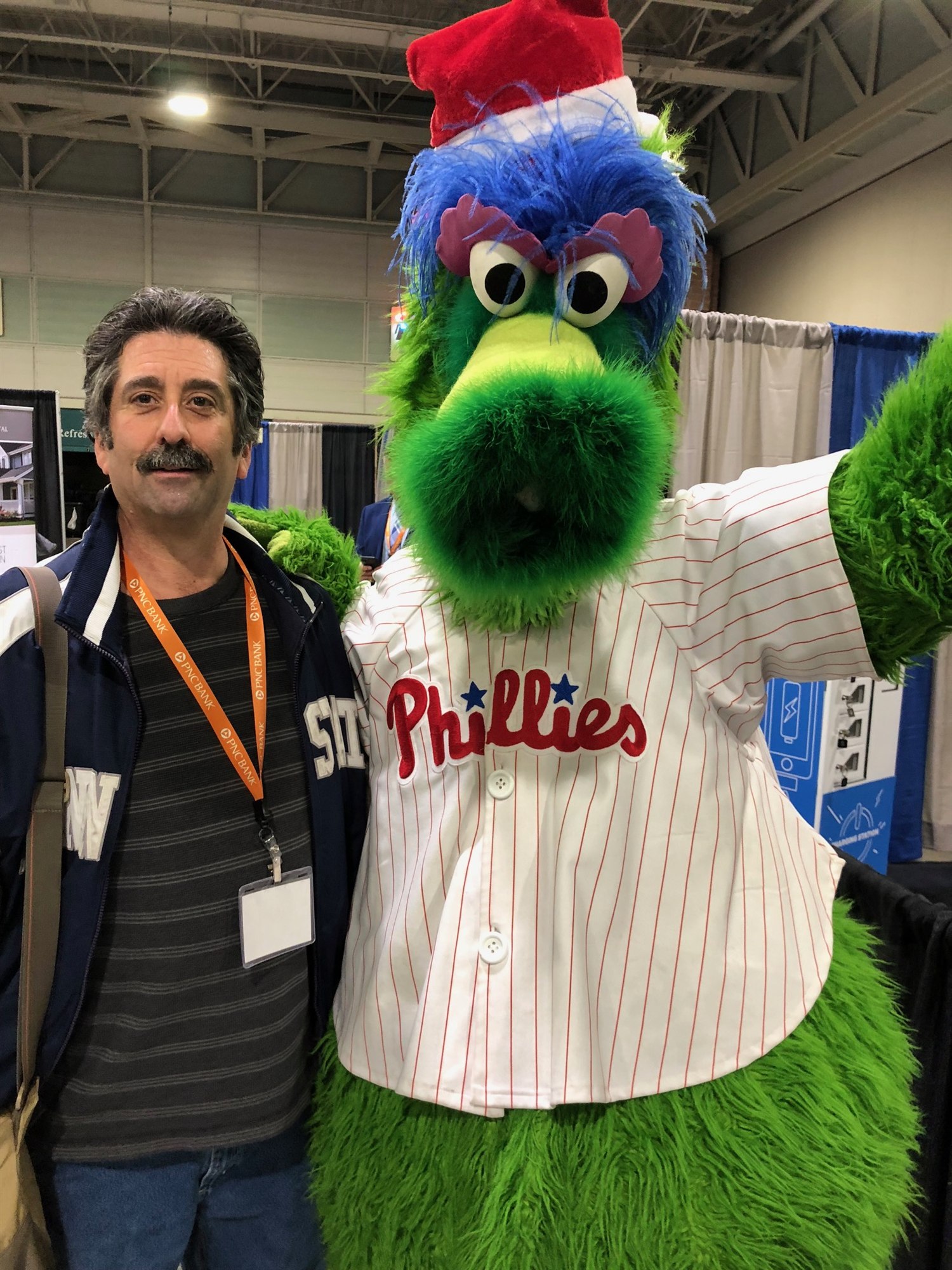 A man in a pinstripe shirt with a convention badge poses on the expo floor with the Phillie Phanatic mascot in a baseball jersey.