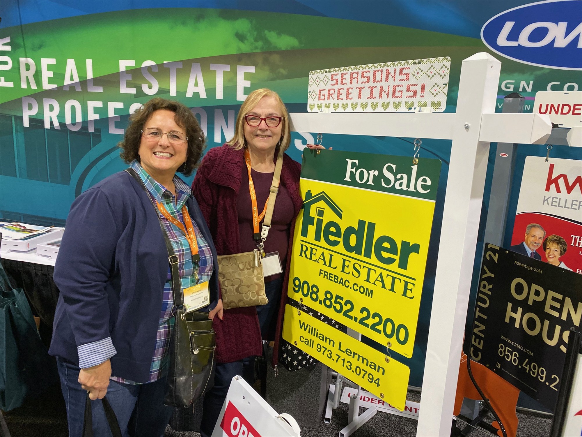 Two smiling women with convention lanyards stand beside a yellow Fiedler Real Estate for-sale sign bearing William Lerman's name, at a sign company's expo booth.