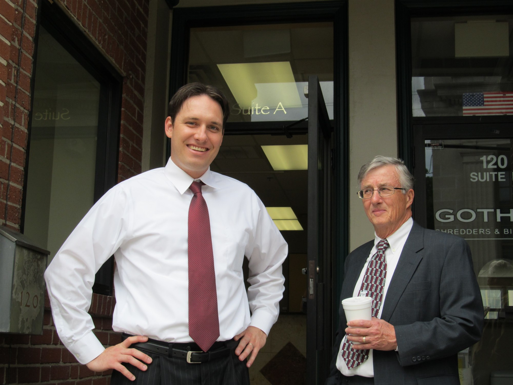 A younger Jeff Fiedler in shirt and tie stands with Henry Riewerts, gray-haired in a suit with a coffee cup, outside an office doorway marked Suite A.