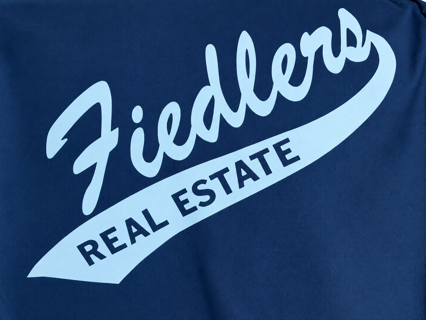 The back of a navy Fiedler championship jacket, printed with Fiedlers in pale-blue script above a swooping banner reading Real Estate.