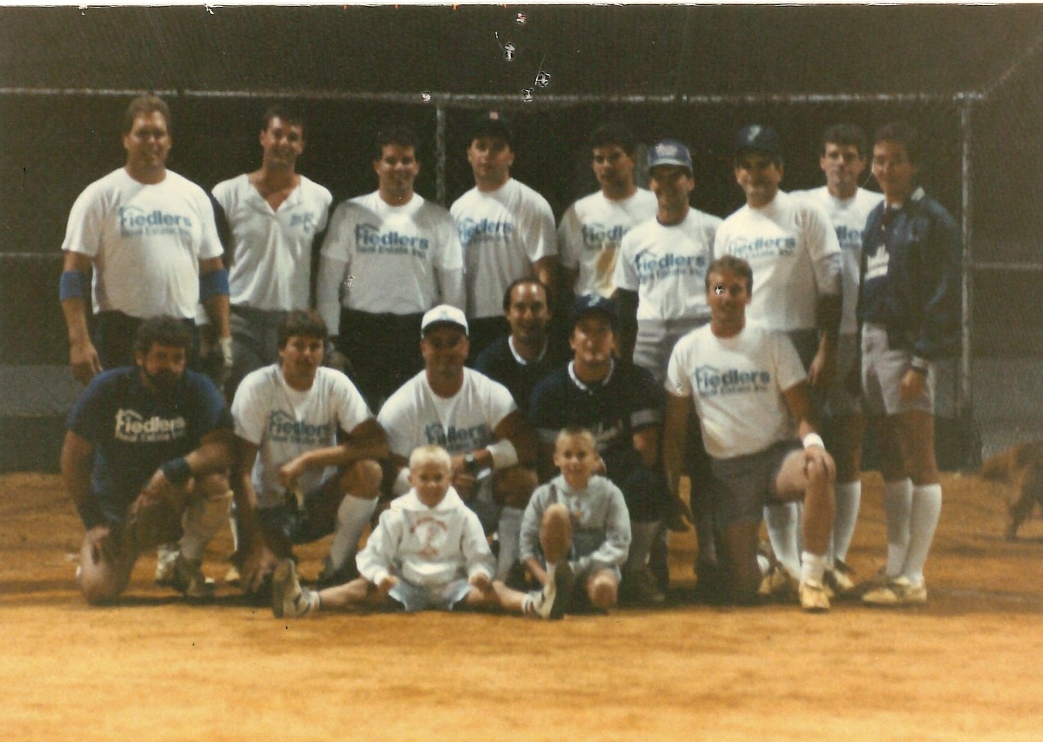 A color team photograph on a lit ballfield: thirteen men in Fiedlers Real Estate shirts in two rows, two small blond boys sitting cross-legged on the dirt in front of them.