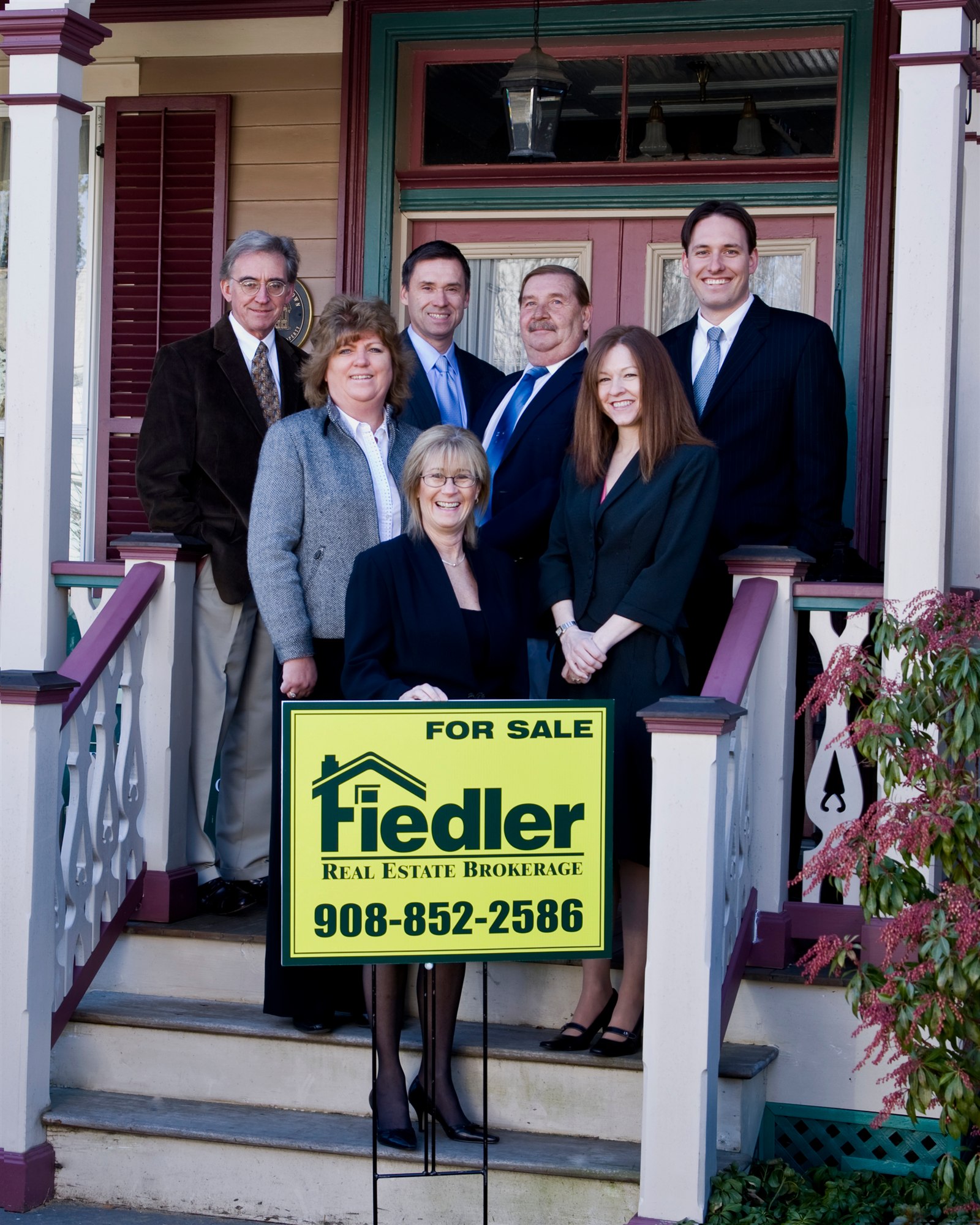 Seven Fiedler agents in business dress gathered on the Victorian porch of the office, behind a yellow Fiedler Real Estate for-sale sign on the steps.