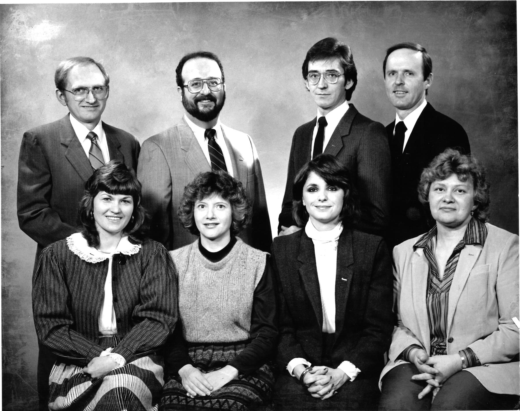 A studio group portrait of the Fiedler Real Estate staff in the mid-1980s — four people standing and four seated.