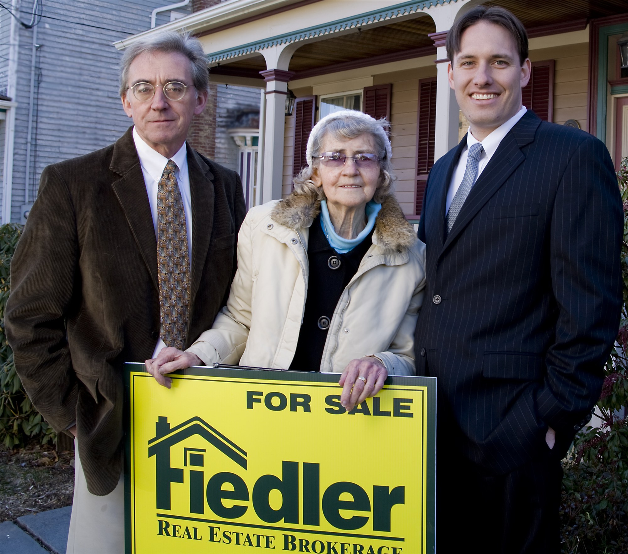 Three people behind a yellow Fiedler Real Estate for-sale sign on a lawn: an older man in a corduroy jacket, an older woman in a cream coat and white hat, and a younger man in a pinstripe suit.