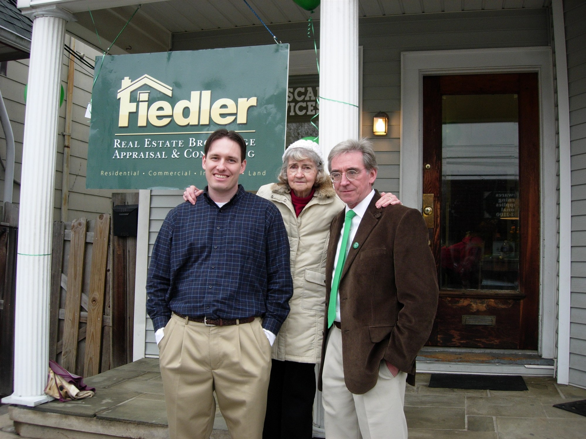 The same three family members on the porch of the Fiedler office at 227 Main Street, beneath the green Fiedler Real Estate Brokerage sign, with green balloons and ribbon on the columns.