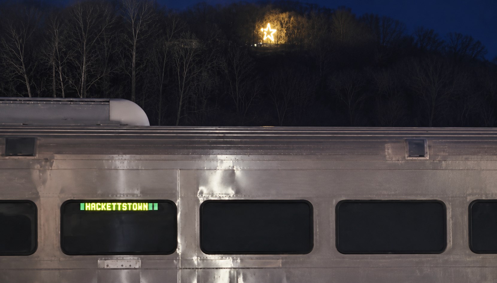 The lit Hackettstown Christmas Star glowing on a wooded hillside at dusk, above a silver commuter train whose lighted sign reads HACKETTSTOWN.