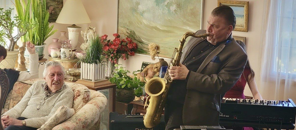 Ed Coyne, in his late nineties, seated in a floral armchair in his living room, smiling as a saxophonist in a gray suit plays tenor sax beside him.