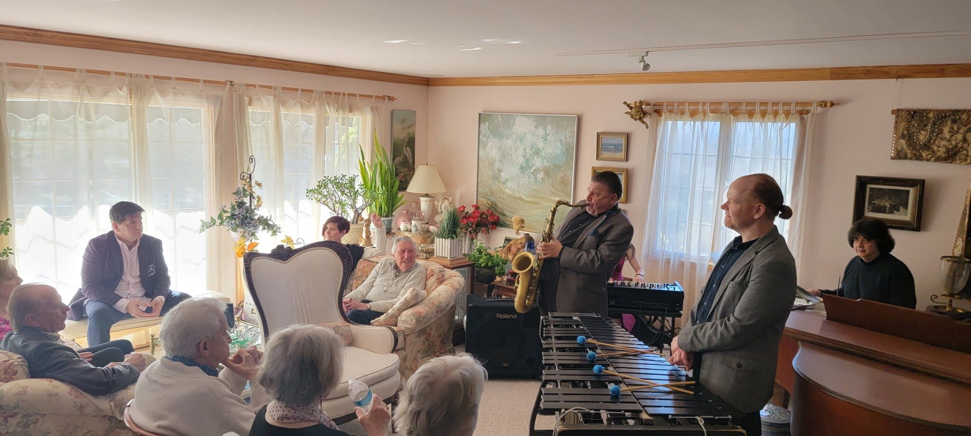 A living-room jazz concert: a saxophonist and a vibraphone player perform for Ed Coyne and a room full of friends seated on couches.