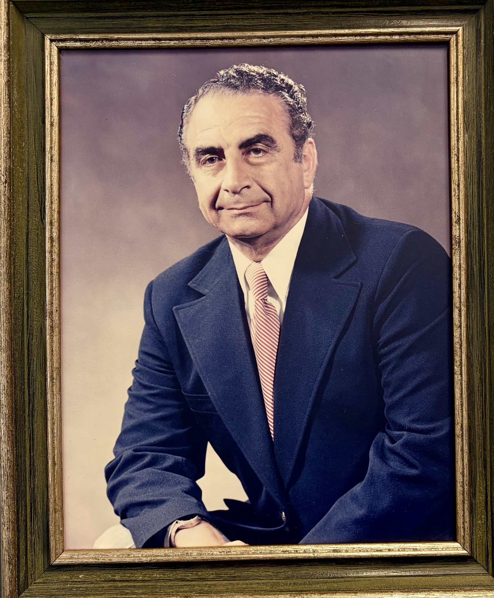 A framed studio portrait of Jacob Garabed in a navy blazer and red-striped tie, dark curly hair, a patient half-smile.