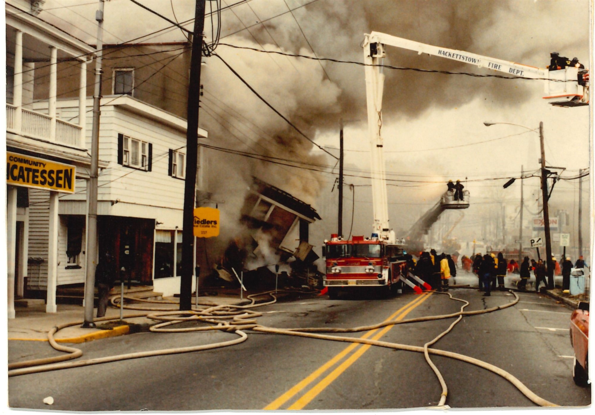 A fire engine and two tower ladders battle a smoking building collapse on Main Street, hoses crossing the road, with a yellow Fiedlers Real Estate sign standing beside the fallen storefront.