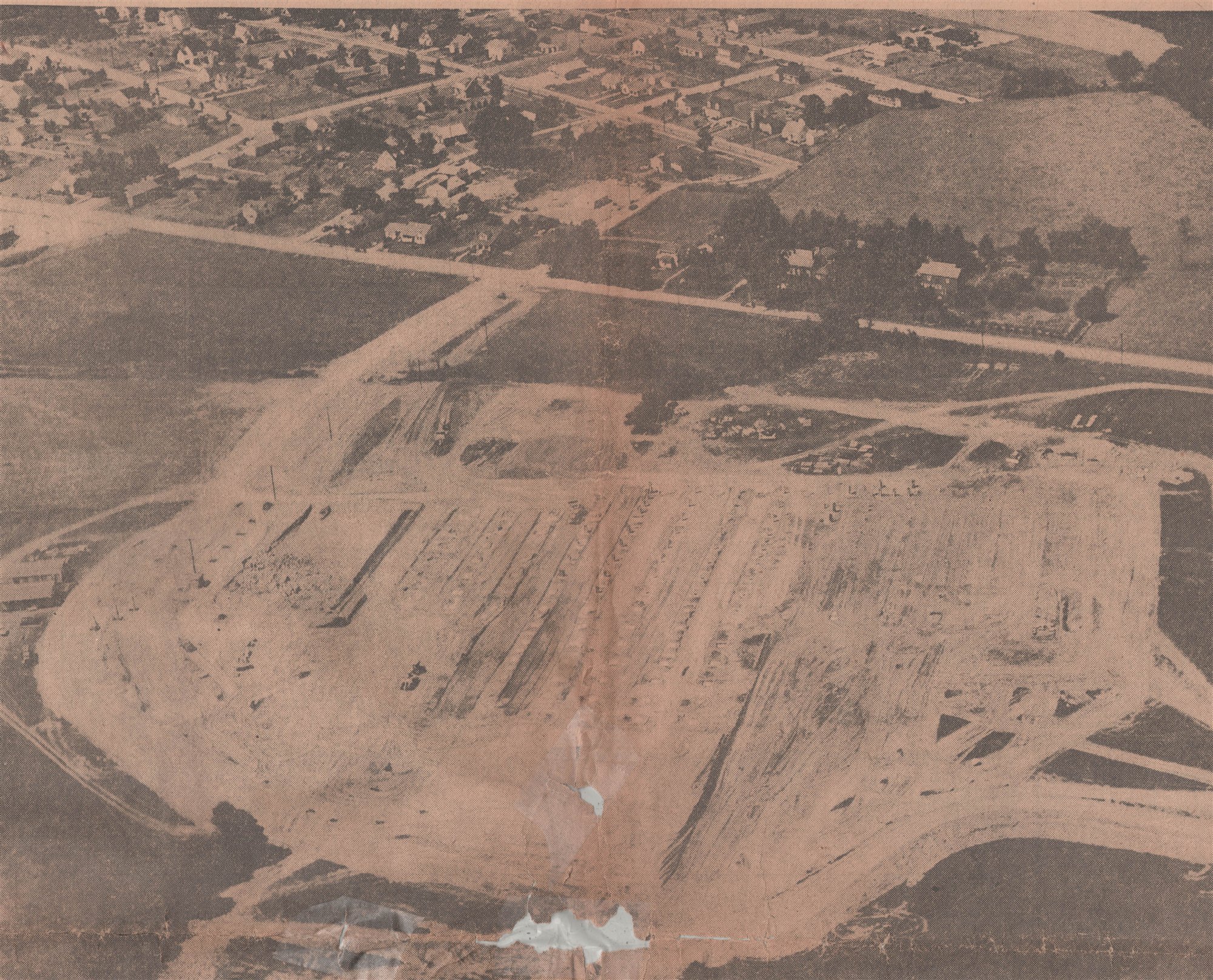 A 1956 aerial photograph from the Hackettstown Gazette: the town's houses at top, and below them the cleared earth of the former Bakker farm, graded rows and footings where the M&M plant is rising.