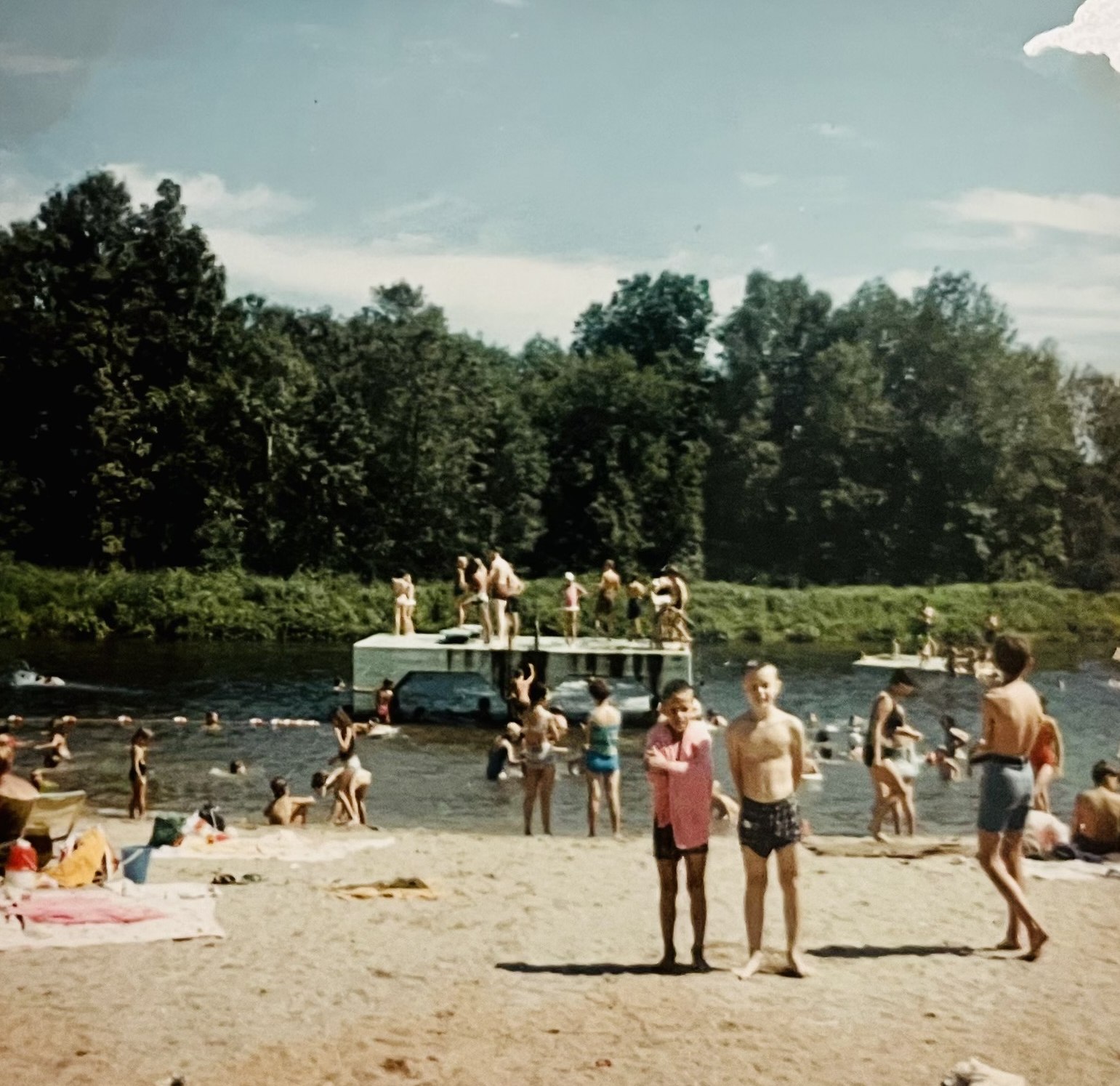 Color photograph of a crowded summer day: two boys pose on the sand, one wrapped in a pink towel, while swimmers fill the roped swim area and children stand atop the white float.