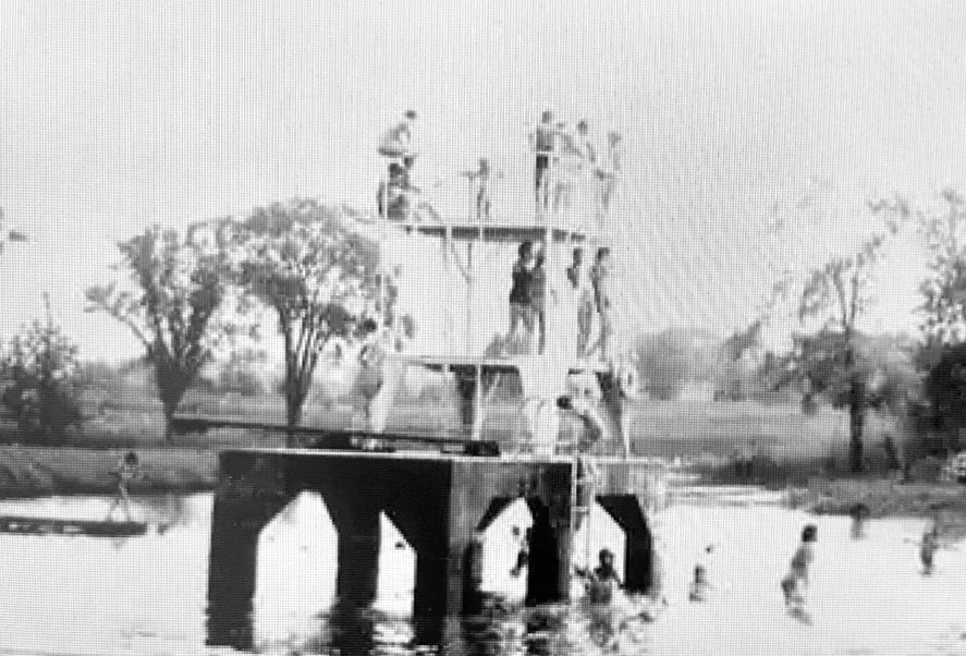 The Sandbar Beach's tall wooden diving tower on a concrete pier in the river, children climbing its levels and swimmers in the water below.