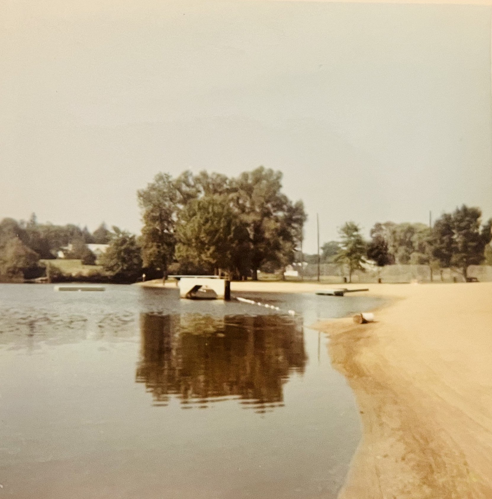 Faded color photograph of the empty sandy beach curving along calm water, floats offshore, and tennis-court fences visible through the trees.