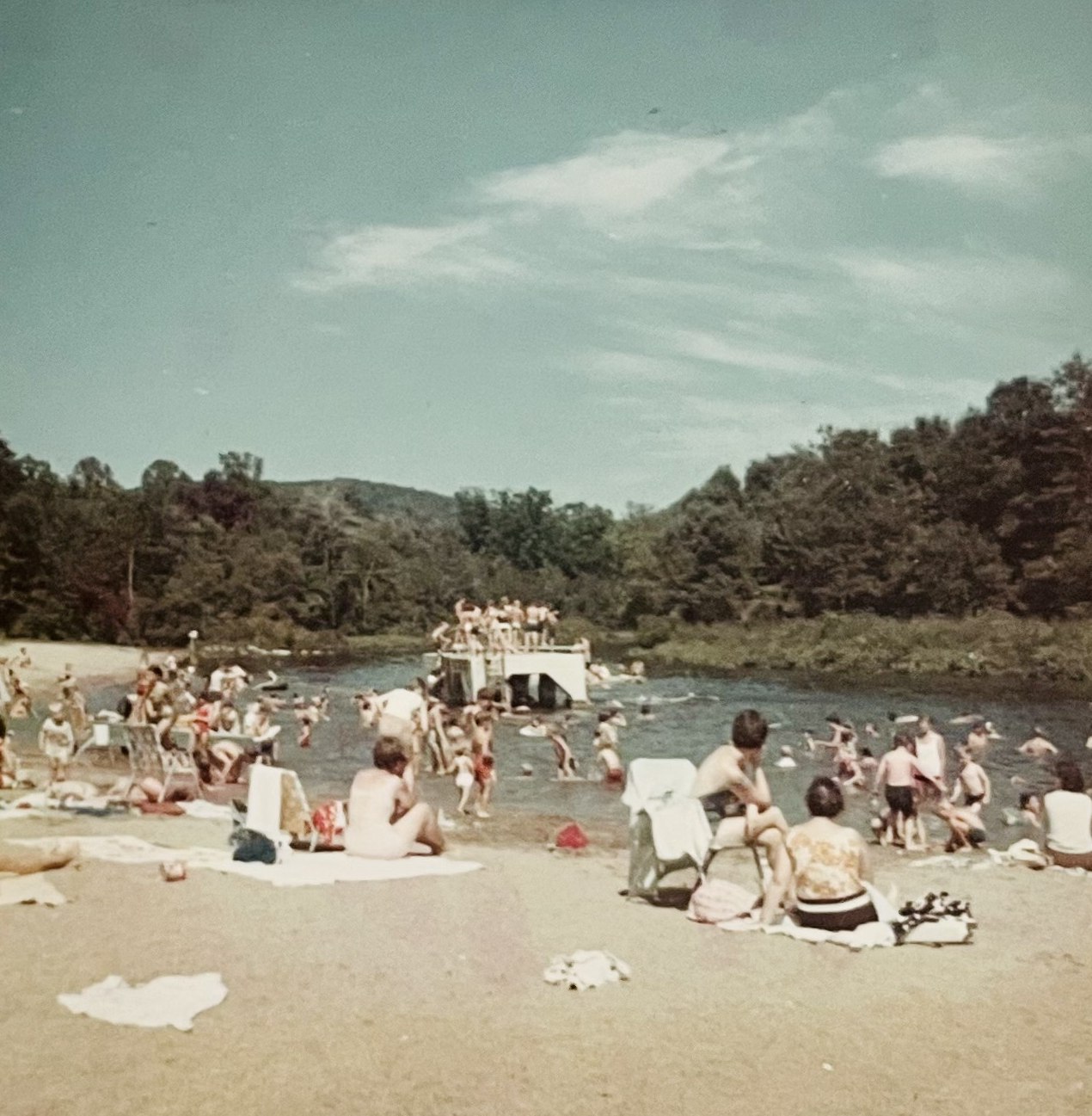 Color photograph of families on beach blankets and folding chairs along the sand, dozens of swimmers in the river, and the swim float busy with children, wooded hills behind.