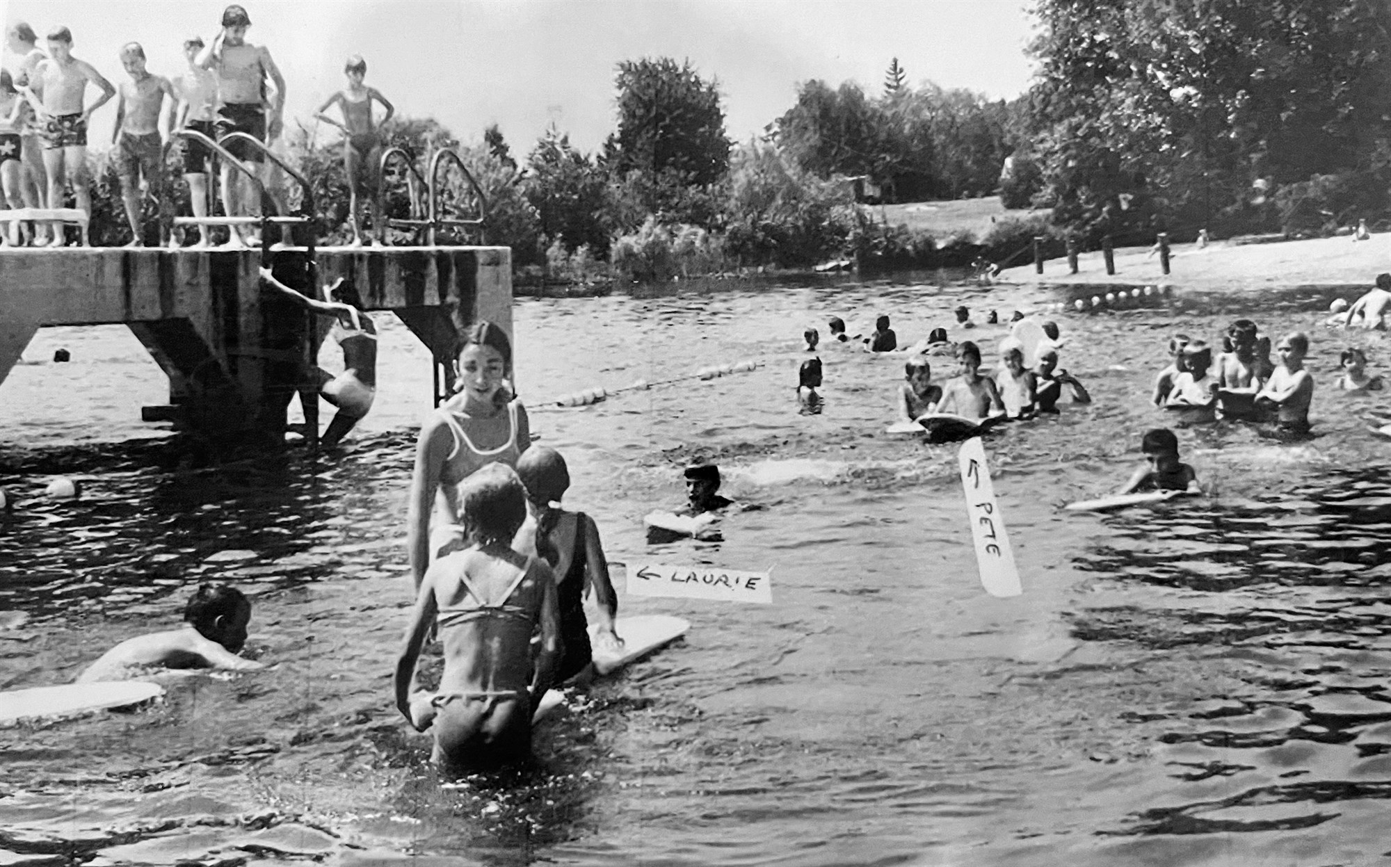 Black-and-white river-beach scene: a young woman in a one-piece bathing suit stands among children holding kickboards, more children lined up on a diving platform at left.