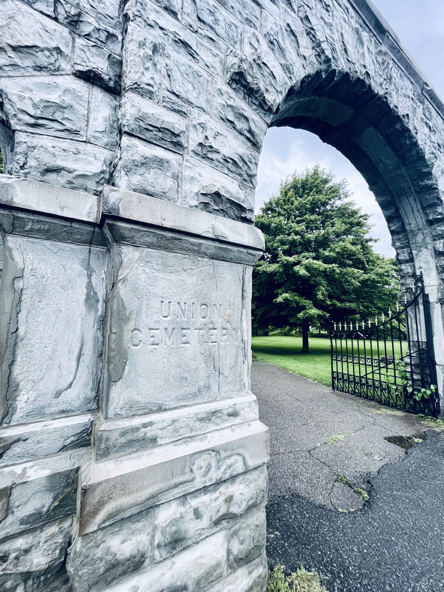 A close view of the cemetery's stone arch with the words 'UNION CEMETERY' carved into the rough gray stone, an open iron gate and green lawn beyond.