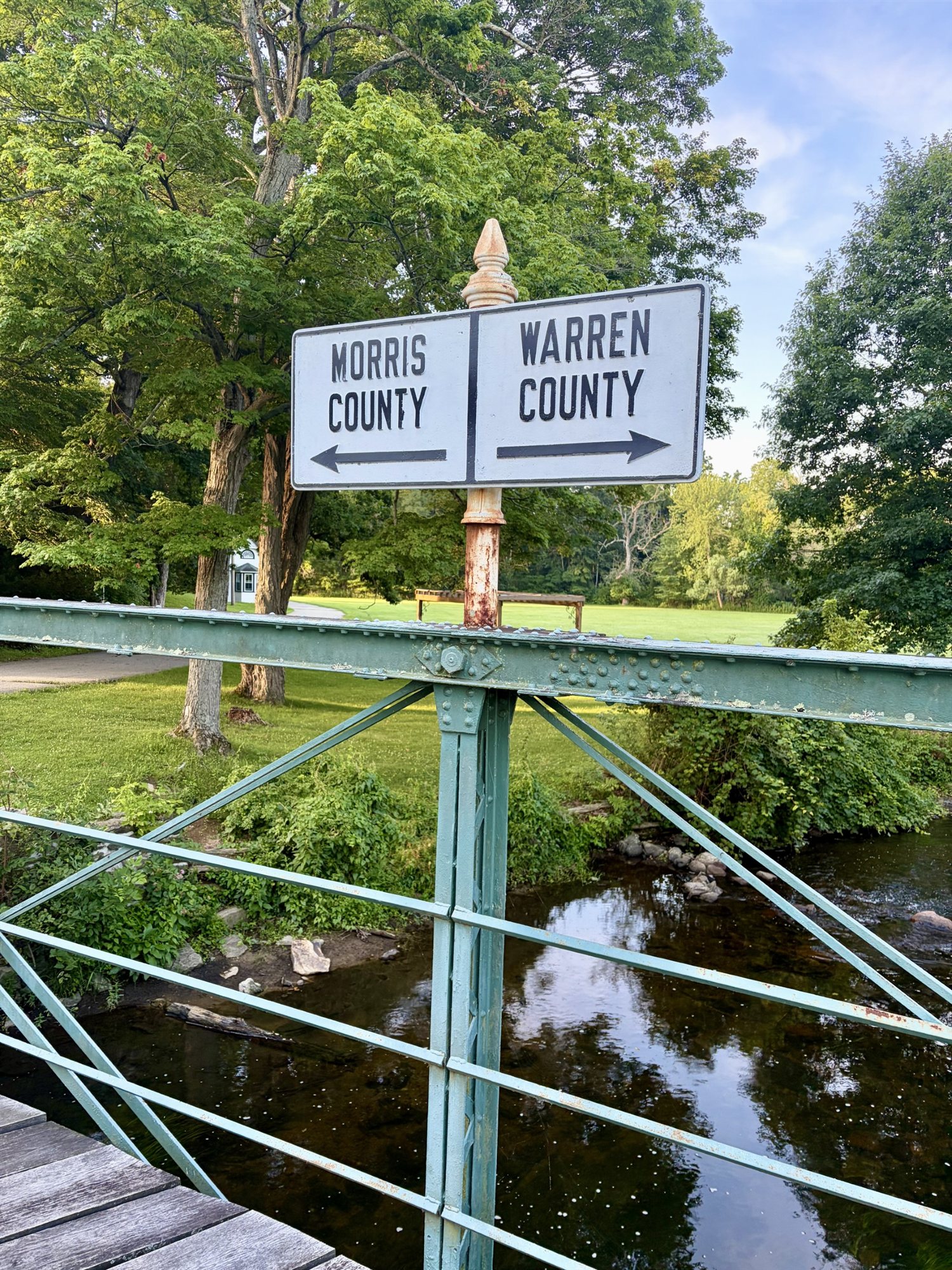 A pale-green iron truss bridge over the Musconetcong River, topped by a vintage sign with arrows pointing left to Morris County and right to Warren County.