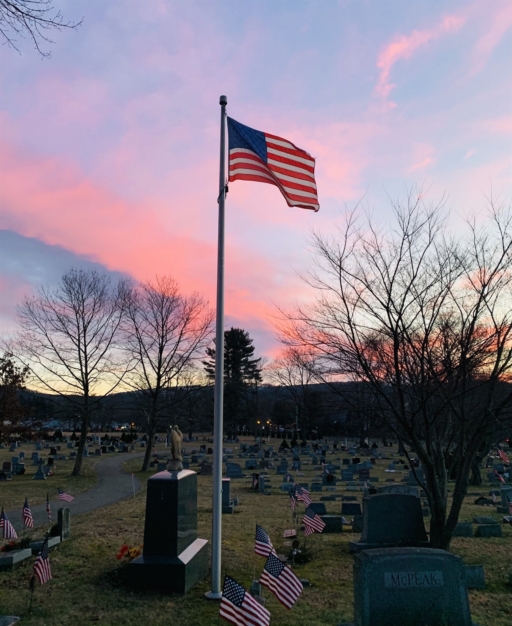 The American flag on a tall pole at the center of Union Cemetery at sunset, rows of gravestones with small flags below and wooded hills glowing pink behind.