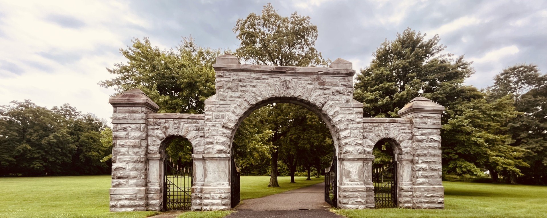 A wide rough-cut stone archway with a central carriage arch and two smaller iron side gates, opening onto a tree-lined cemetery drive under a cloudy sky.