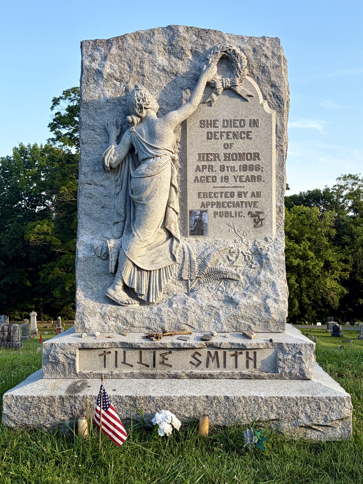 A granite monument carved with a robed woman lifting a wreath, inscribed 'She died in defence of her honor,' the base lettered 'TILLIE SMITH,' with a flag and flowers at its foot.
