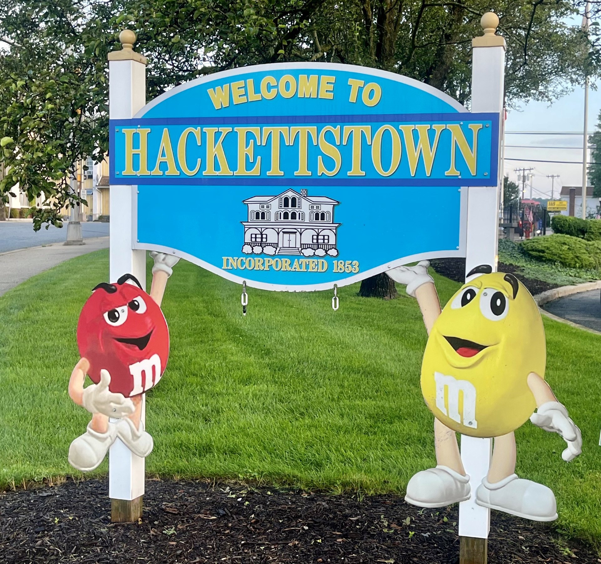 A blue 'Welcome to Hackettstown, Incorporated 1853' sign with a Victorian-building emblem, held up by red and yellow M&M character cutouts on a green lawn.