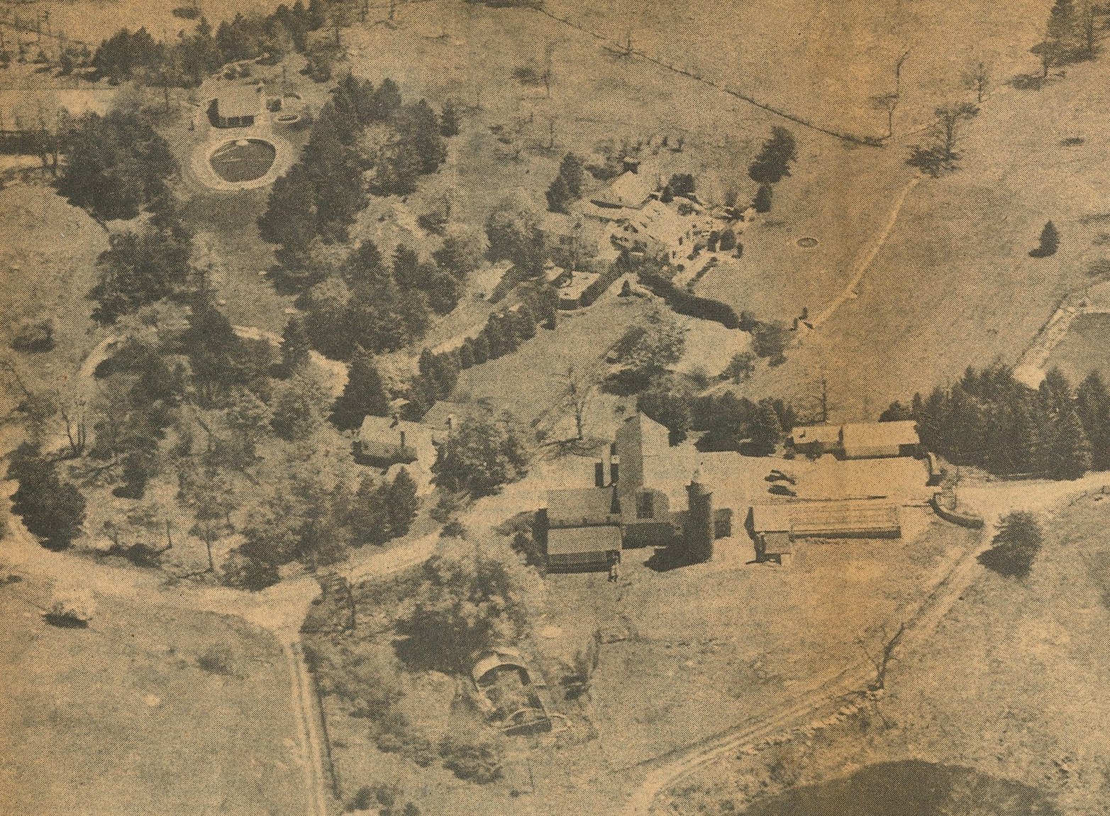 A 1959 aerial view of the Panther Ledge estate in Allamuchy: the Colonial mansion among trees, barns and outbuildings along dirt lanes, the oval pool at upper left, open fields beyond.